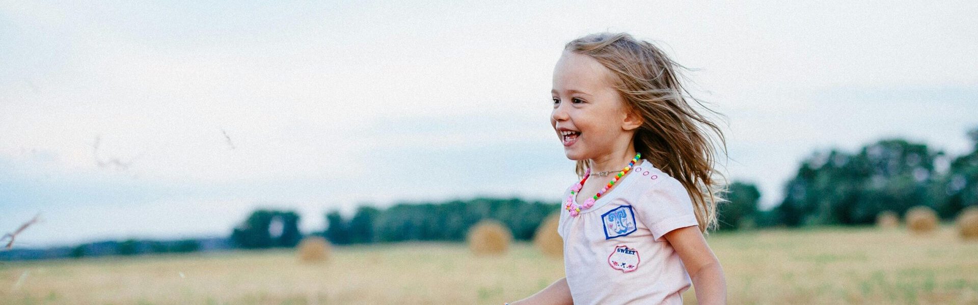 A happy female child is running toward someone off-camera in a field, with round bales in the distance.