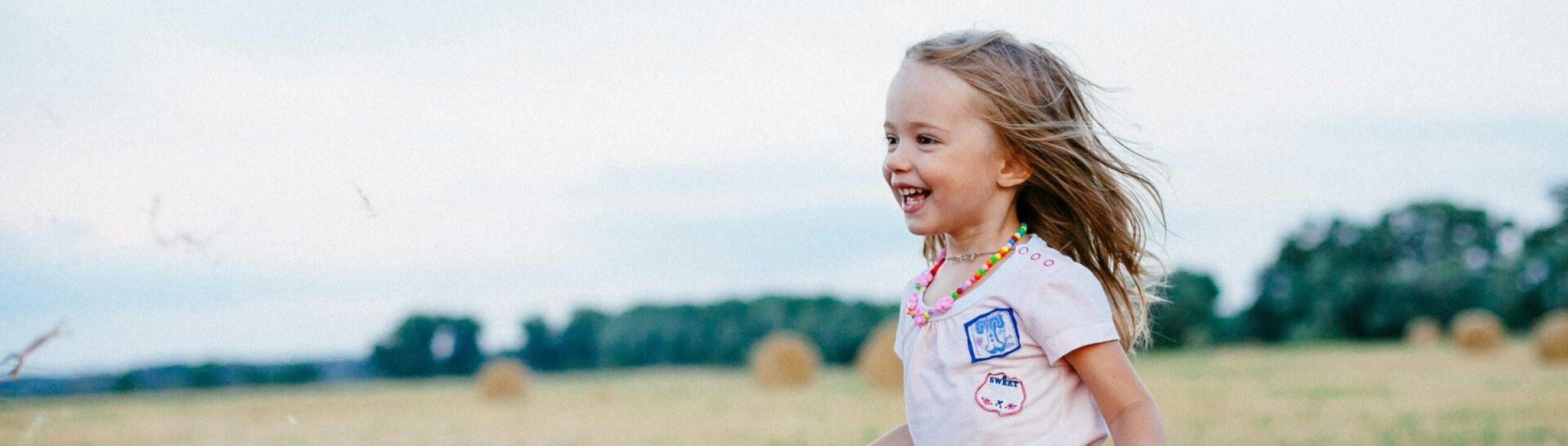 A happy female child is running toward someone off-camera in a field, with round bales in the distance.